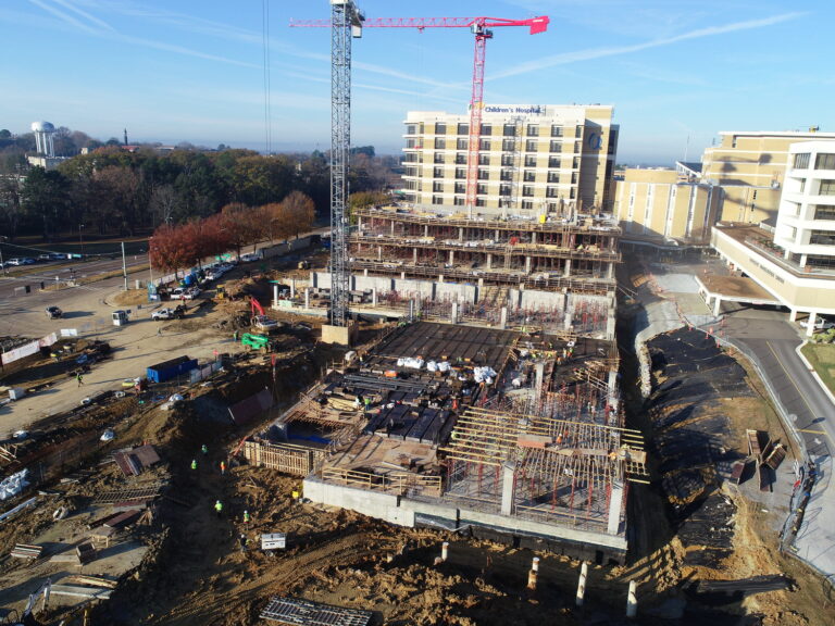 Aerial view of a construction site with a multi-story parking deck under development by Brasfield & Gorrie, featuring scaffolding, a crane, and construction materials, adjacent to a roadway.