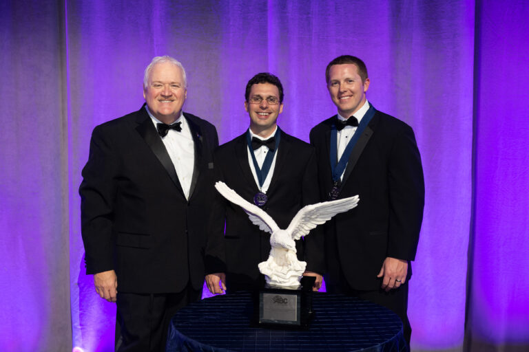 Three men in formal attire posing with an eagle statue award at an event.