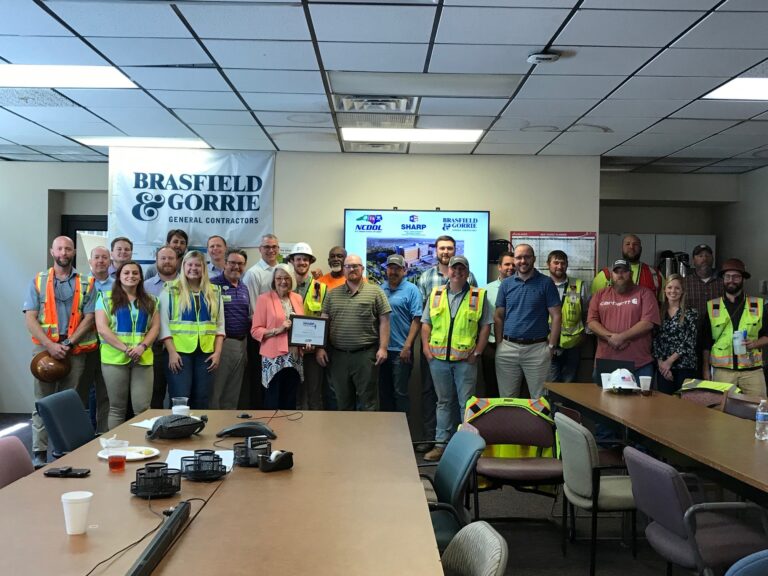 Group of construction workers in safety vests gathered for a meeting or event inside an office setting.