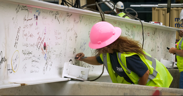 Construction worker in a pink hard hat signing a beam surrounded by other signatures.