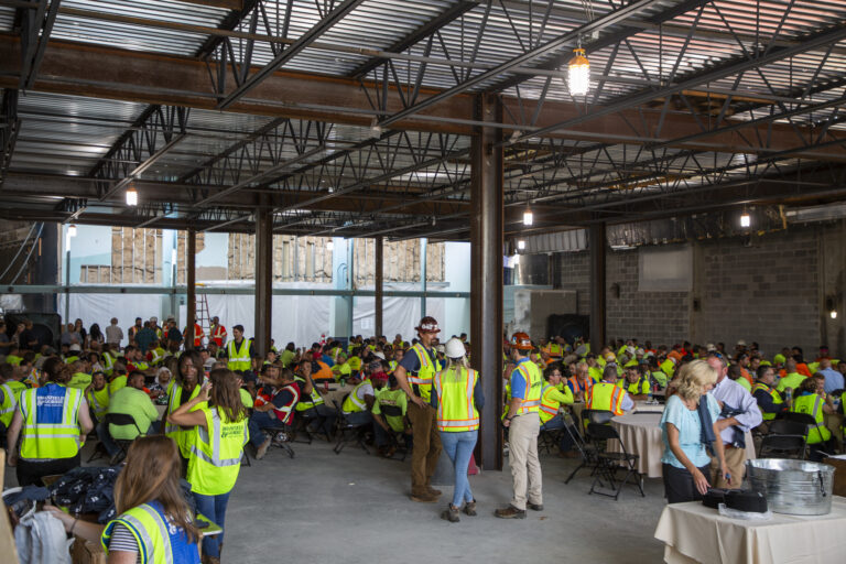 Workers in high-visibility vests gathered in an industrial building under construction for a meeting or event.