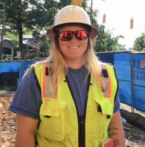 A smiling woman wearing a hard hat, reflective safety vest, and sunglasses at a construction site.