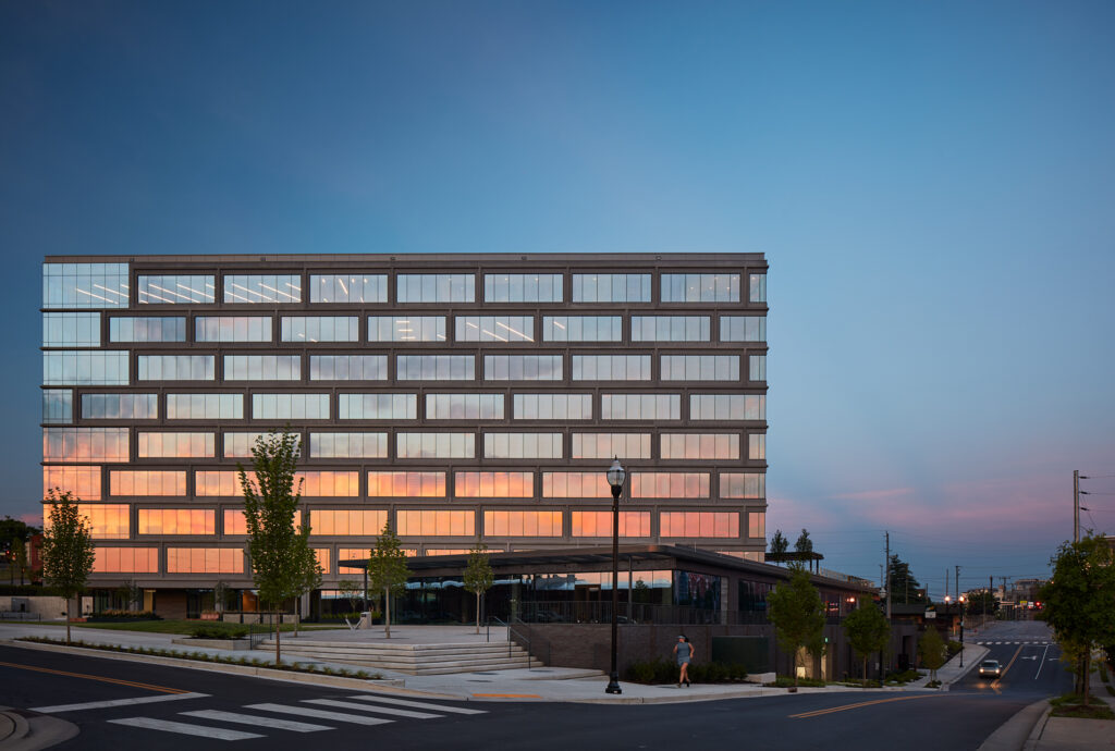 Modern office building at twilight with illuminated windows and a pedestrian walking by.