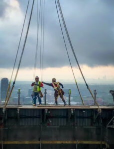 Construction workers reaching new heights at a high elevation on a building site, secured with safety harnesses, against a backdrop of a cloudy sky and city skyline.