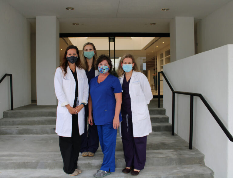 Four healthcare workers wearing face masks and lab coats are standing on steps outside the Alabama Fertility Specialists facility, celebrating its completion.