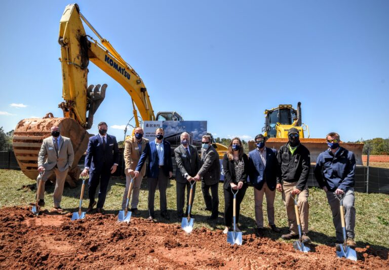 Group of individuals in formal attire participating in a groundbreaking ceremony with shovels for the Greenville Data Center, while construction equipment is visible in the background.