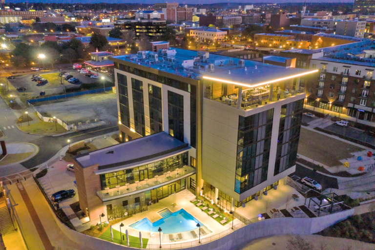 Aerial view of the Hotel Indigo in Columbus, Georgia, featuring a modern building with a rooftop pool illuminated at dusk, surrounded by urban development.