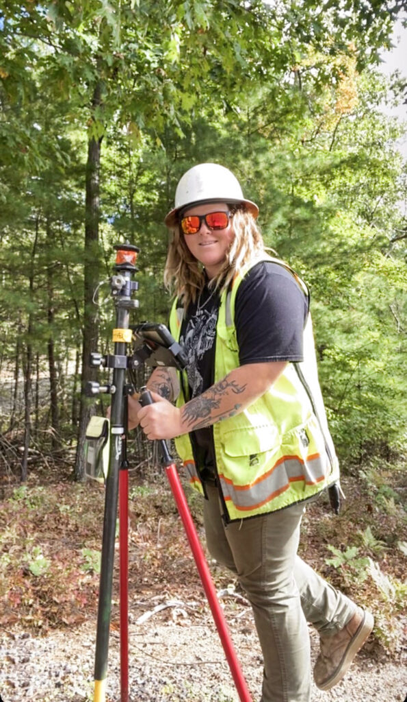 A surveyor in high-visibility gear operates equipment in a wooded area