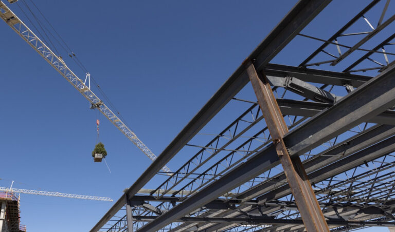 A crane lifting materials above a steel structure under construction against a clear blue sky for the Omni PGA Frisco Resort.