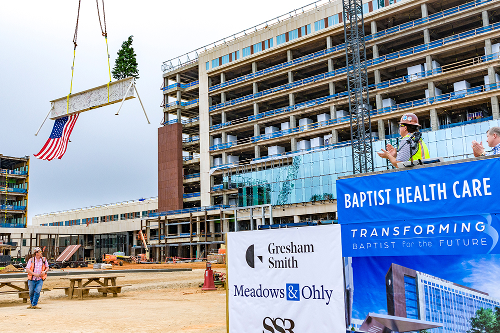 Construction workers observing as a large American flag is hoisted at a building site with a crane during the Topping Out ceremony organized by Brasfield & Gorrie for the Baptist Health Care project.