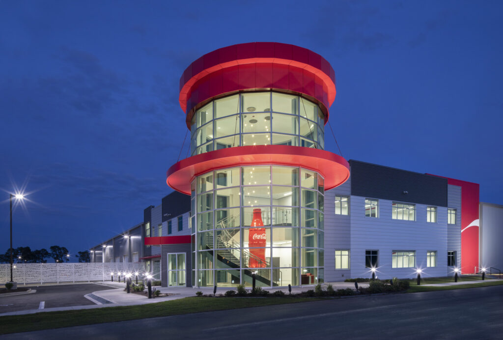 Modern building with a distinctive red cylindrical tower illuminated at twilight opens as Coca-Cola UNITED Tifton Sales Center.