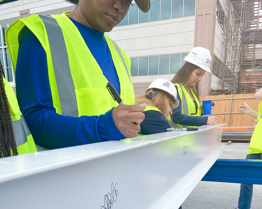 Construction workers in safety gear signing a beam at the North Austin Medical Center construction site.
