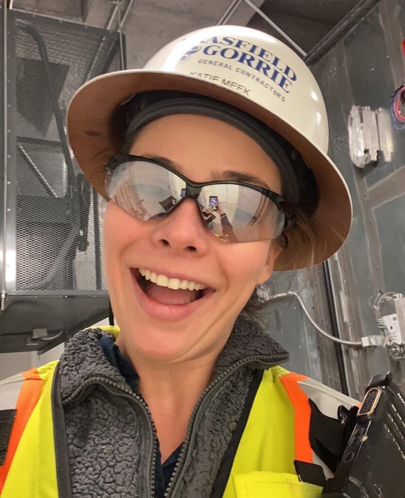 A smiling woman wearing a hard hat, safety glasses, and a high-visibility vest on a construction site during Construction Week.