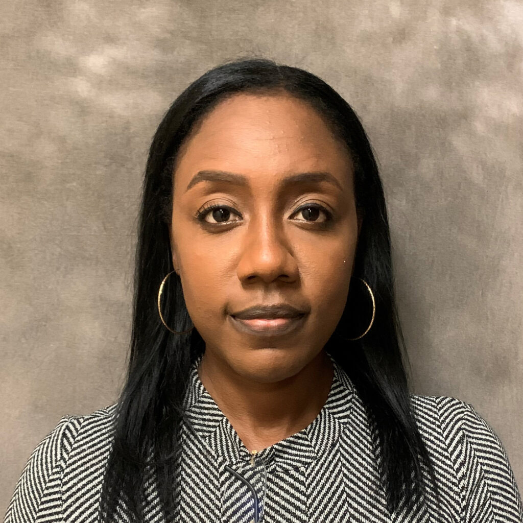 A woman with hoop earrings and a houndstooth top poses for a portrait against a gray background, symbolizing progress during Women in Construction Week.