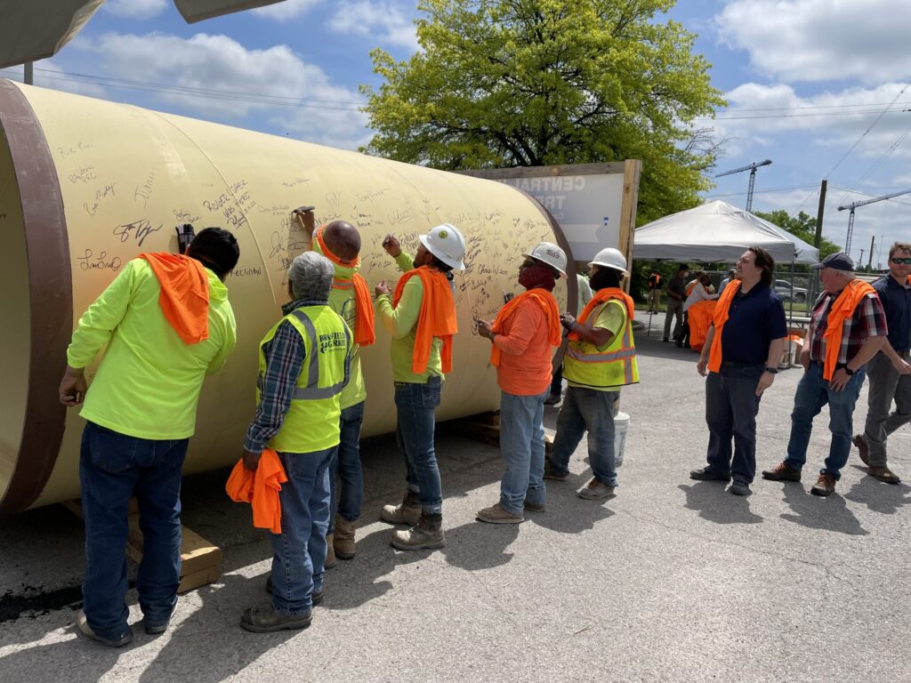 Group of construction workers in safety vests and helmets signing a large cylindrical structure at the Central Wastewater Treatment Plant.