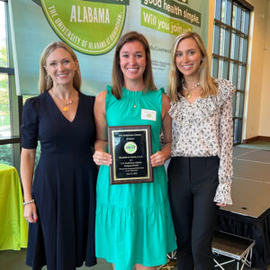 Three women smiling at an award ceremony, with one holding a commemorative plaque for their designation in Live HealthSmart Alabama.