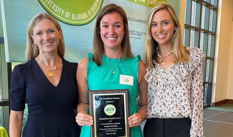 Three women smiling, with the center woman holding an award plaque for her designation by Live HealthSmart Alabama.