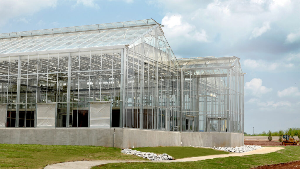 Modern greenhouse under construction with a clear framework and partial cladding, set against a partly cloudy sky, scheduled for HudsonAlpha's Greenhouse opening.