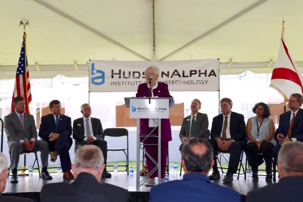 A person speaking at a podium during the Greenhouse Opening event while several individuals sit on stage, with flags and HudsonAlpha organizational banners in the background.