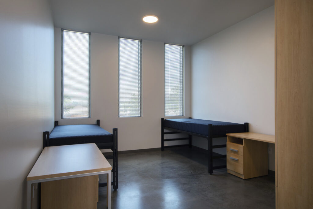 A minimalist dormitory room at the Alabama School of Cyber Technology and Engineering, designed for the 2022-2023 school year, features two beds, desks, and windows with blinds.