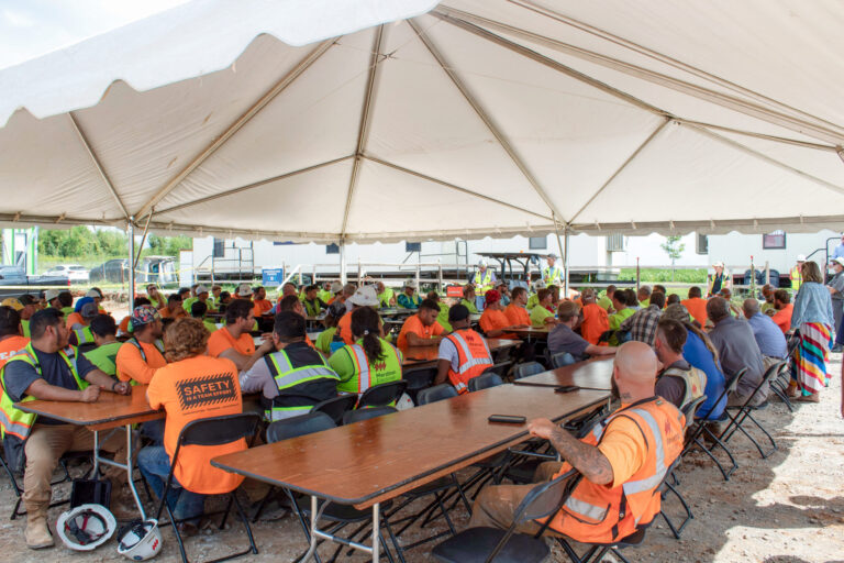 Workers in high-visibility vests attending a Tops Out meeting for the Brasfield & Gorrie Life Sciences Facility under a large tent.