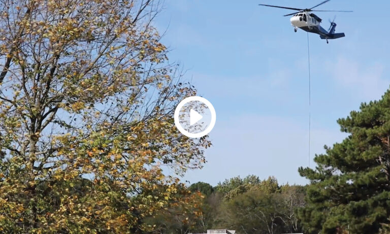 A helicopter performing an HVAC installation hovers near trees with a visible cable hanging from its underside.