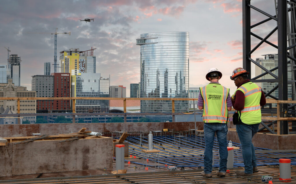 Two construction workers with reflective vests and hard hats are standing at a construction site, engaged in self-perform concrete tasks, with a city skyline in the background.