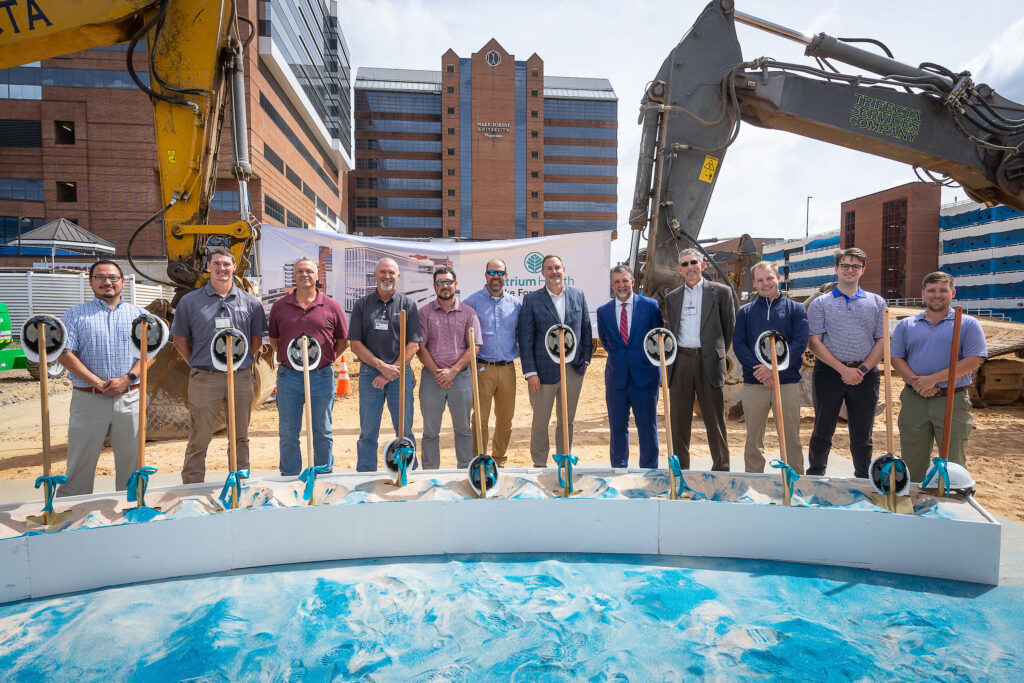 Group of individuals in business and construction attire participating in a groundbreaking ceremony for the New Care Tower at Atrium Health with shovels and hard hats.
