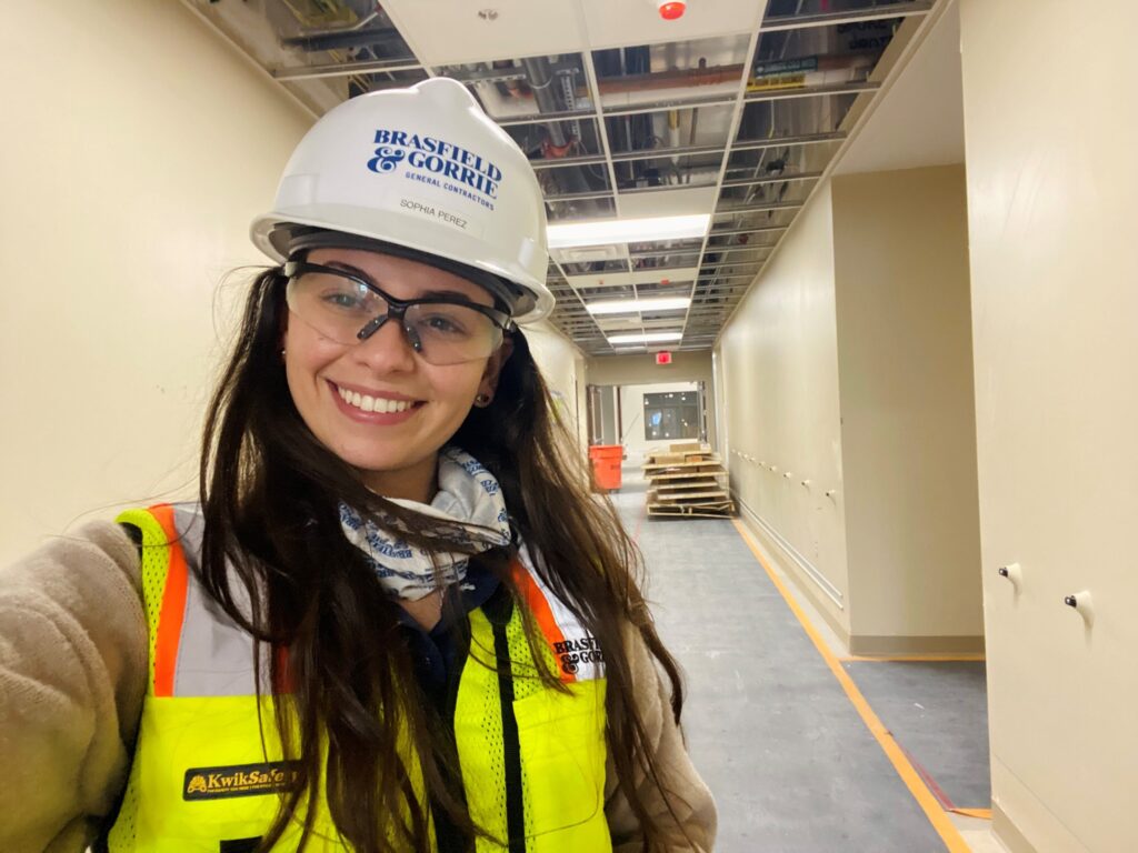 A woman in a hard hat and safety vest takes a selfie in a construction site corridor.