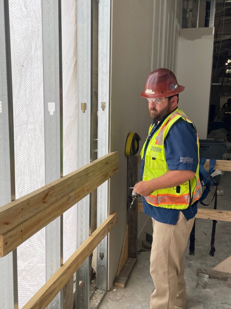 A construction worker wearing a hard hat and safety vest standing by a metal door frame on a construction site.