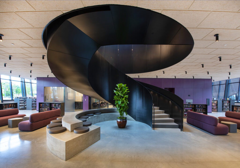 Modern library interior featuring a spiral staircase with black balustrades, surrounded by shelves of books and contemporary seating areas, honored in ENR Projects.