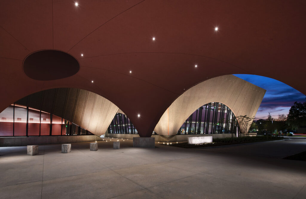 An architectural photograph capturing one of the Four Projects, showcasing a modern building with an arched, cantilevered canopy and curved glass facades against a dusk sky.