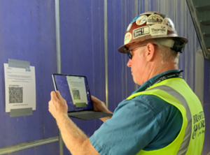 A worker in a hard hat and reflective vest scans a QR code with a tablet for digital dig boards.