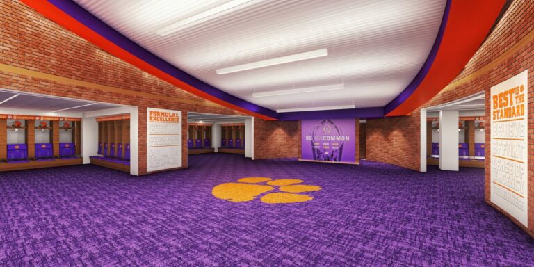 An empty sports locker room with orange and purple lockers, vibrant purple carpet, and team banners on the walls is part of the second phase of Clemson Football Stadium renovations.