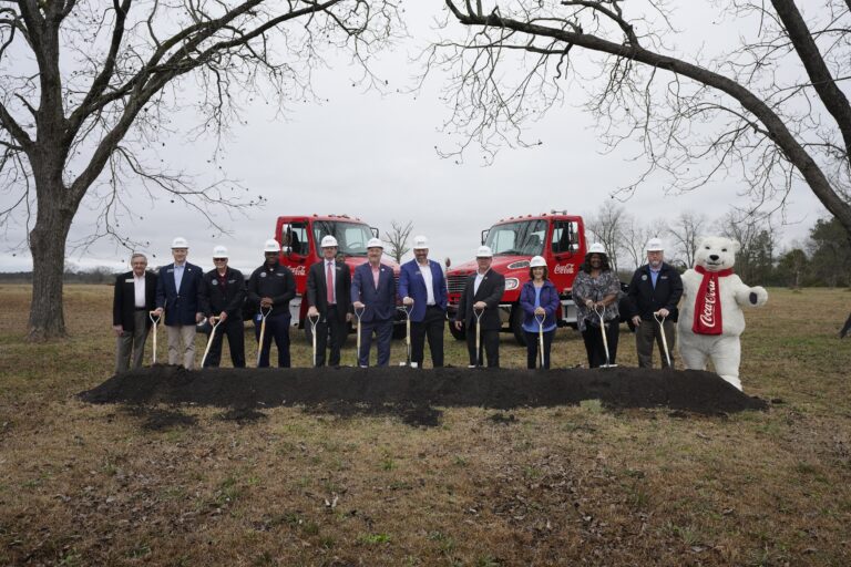 A group of individuals with shovels at a ground breaking event for the Beverage Facility Expansion, flanked by Coca-Cola branded vehicles and a Coca-Cola mascot.