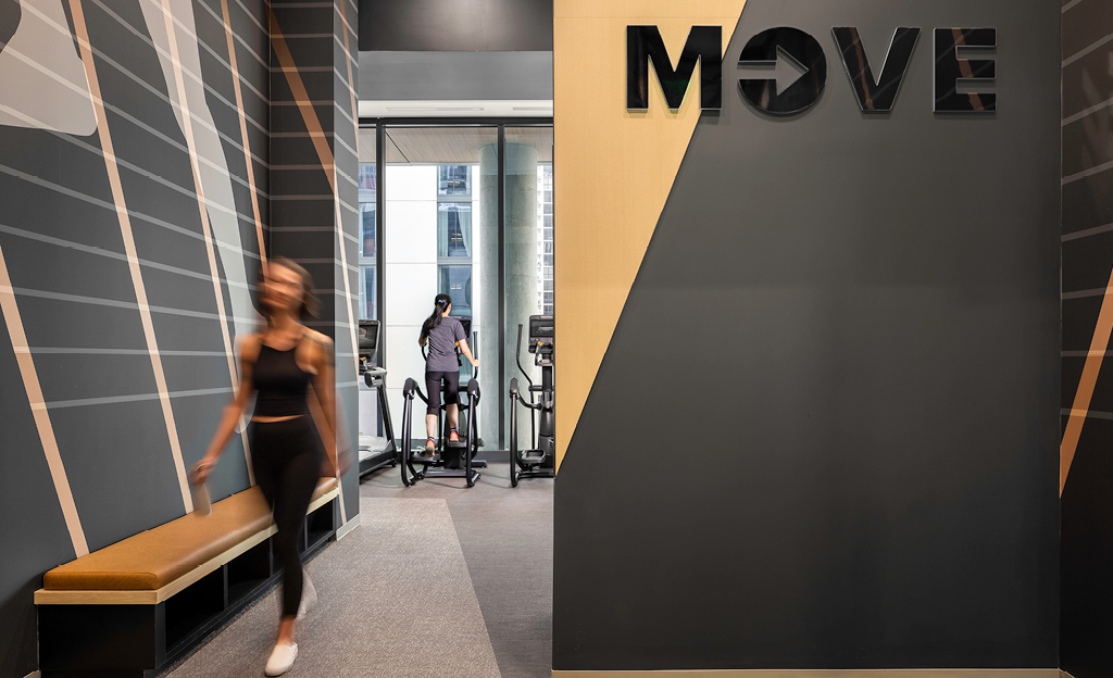 Woman walking past a gym area in Atlanta's Midtown Union with the word "move" on the wall, while another person exercises on an elliptical machine inside.