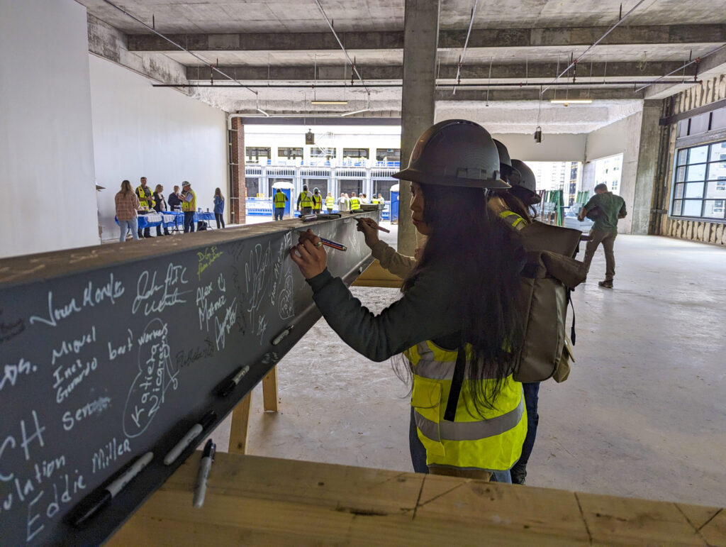 A person in a hard hat and reflective vest writing "Phase One, 2024 Opening" on a blackboard at a construction site on High Street.