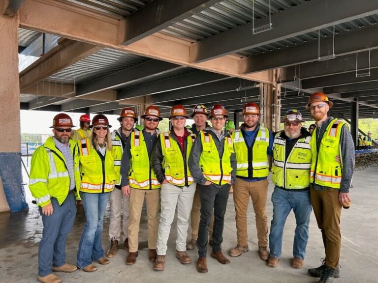 A group of construction workers in hard hats and high-visibility vests posing together at a biomanufacturing campus construction site.