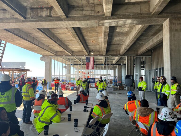 Construction workers gathered for a meeting under an unfinished concrete structure at a Brasfield & Gorrie manufacturing facility, celebrating a construction milestone with an American flag hanging in the background.