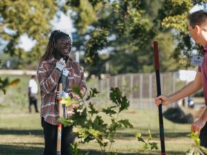 Two people, part of the Brasfield & Gorrie team, are planting trees in a park. One person is smiling while holding a shovel. Trees and a fence provide the perfect backdrop to their community-focused efforts.