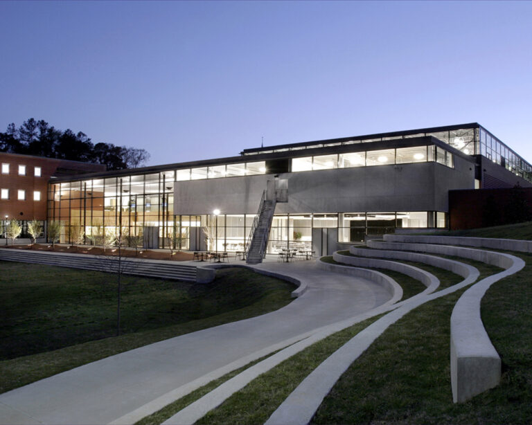 Modern school building with large glass windows illuminated at dusk, featuring terraced landscaping and a curved pathway.