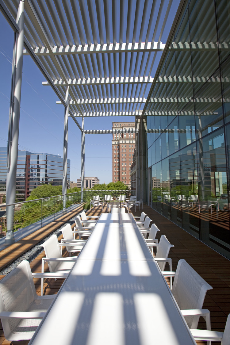 Modern outdoor terrace with a long white table, white chairs, and a louvered roof overlooking an urban landscape.