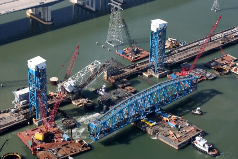 Aerial view of heavy civil construction of a bridge with cranes and barges on a river.