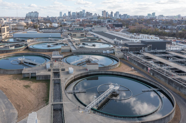 Aerial view of a wastewater treatment plant focusing on heavy/civil construction with circular clarifier tanks in the foreground and a city skyline in the distance.