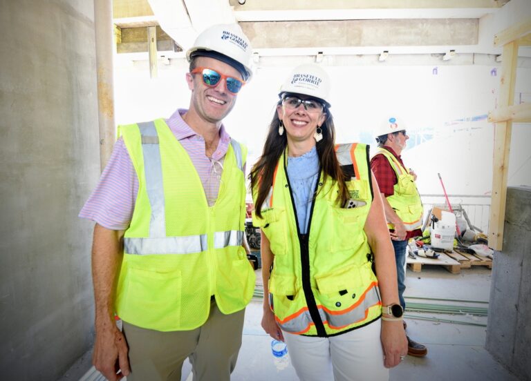 Two people wearing hard hats and high visibility vests smiling on a construction site.