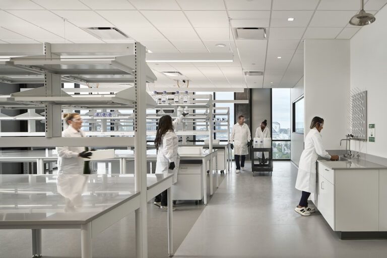 In a Brasfield and Gorrie Science and Technology project, a laboratory bustles with five individuals in white lab coats, each diligently working at various stations surrounded by shelves and tables.