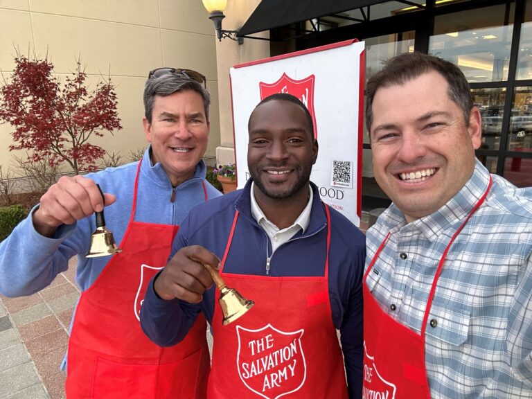 Three individuals volunteering for the Salvation Army in Birmingham, Alabama, holding bells and wearing red aprons, pose for a selfie.