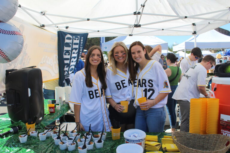 Three smiling individuals in sports jerseys standing at a Birmingham, Alabama promotional event booth with beverages and merchandise on display.