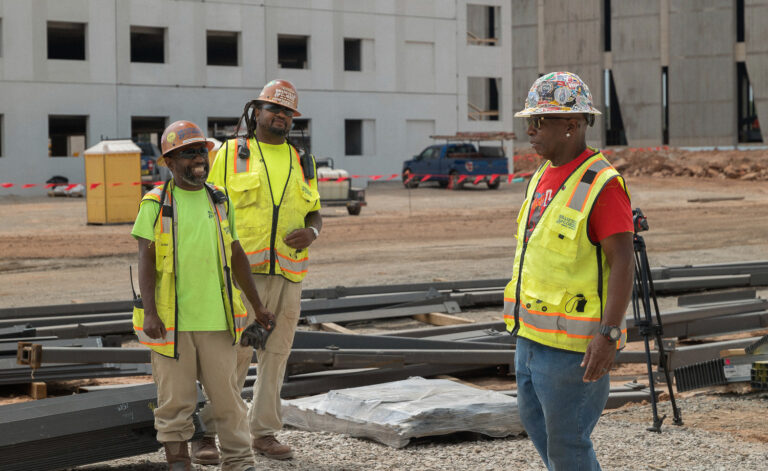 Three construction workers in reflective vests and hard hats discuss on a construction site in Charlotte, North Carolina, with building materials and a structure in the background.