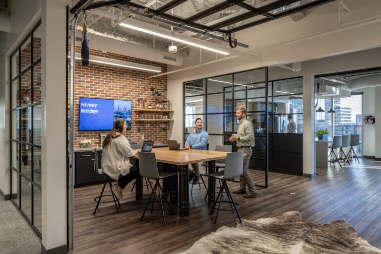 Modern office space in Charlotte, North Carolina, with four people, brick walls, and glass partitions, discussing around a table with laptops near a digital display showing 